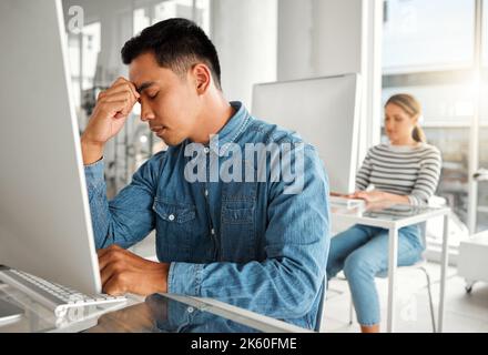 Young stressed asian businessman suffering from a headache while working on a desktop computer at work. One unhappy chinese male businessperson Stock Photo