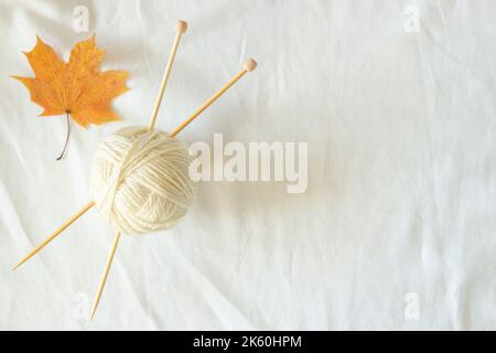 White wool thread ball with wooden bamboo knitting needles and autumn maple fall leaf on white linen fabric background. Top view, copy space. Hobby Stock Photo