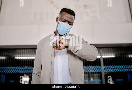African american businessman travelling alone and standing in a train station while checking his travel times on his watch while wearing a mask Stock Photo