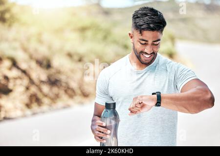 Athletic young mixed race man looking at his watch while exercising outdoors. Handsome hispanic male holding a water bottle and checking the time Stock Photo