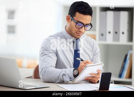 Young focused mixed race businessman writing In a notebook alone in an office at work. One hispanic male businessperson taking notes in a diary Stock Photo