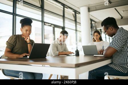 Diverse group of entrepreneurs business people working on laptops and writing notes while sitting around a table in a shared coworking office space Stock Photo