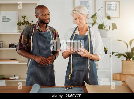 Two designers using a digital tablet while working with material at work. Mature caucasian tailor talking and holding a digital tablet while working Stock Photo