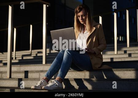 Woman sitting using laptop. Female freelancer working remotely at city street in sunset Stock Photo