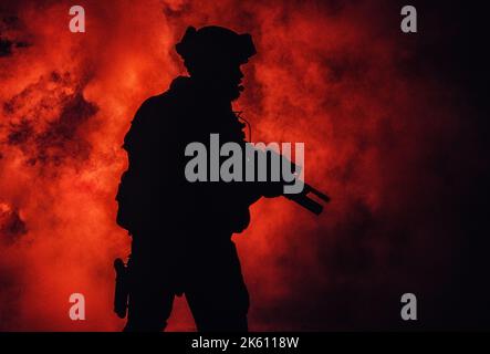 Silhouette of modern infantry soldier, elite army fighter in tactical ammunition and helmet, standing with assault service rifle in hands in the fire and smoke Stock Photo