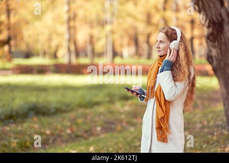 A young red-haired woman in a white coat walks in an autumn park. A girl listens to music in the autumn forest Stock Photo
