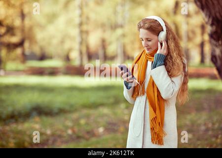 A young red-haired woman in a white coat walks in an autumn park. A girl listens to music in the autumn forest Stock Photo