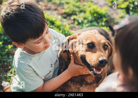 Two caucasian kids playing with their loyal dog, scratching dog's head, caressing, cuddling. Sunny day. Having fun outdoors. Beautiful young girl Stock Photo