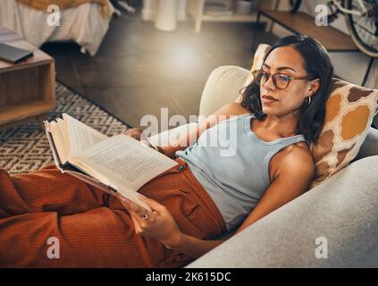 Serious mixed race woman reading a book in living room at home. Beautiful hispanic lying down on lounge sofa alone and enjoying a novel. Relaxed woman Stock Photo