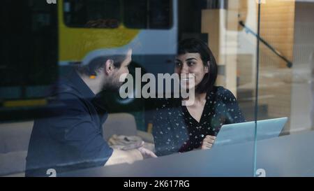 Two young people talking about work in front of laptop and workplace seen through window reflection. Business woman and man discussing in conversation Stock Photo