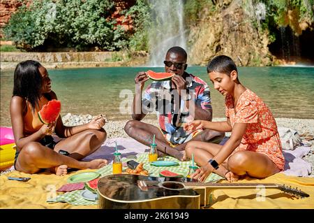 Group of multiracial friends sitting together on plaid and eating watermelon while having picnic against lake in summer Stock Photo