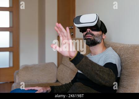 Bearded male in goggles of virtual reality sitting on sofa and raising hand while interacting with another dimension Stock Photo