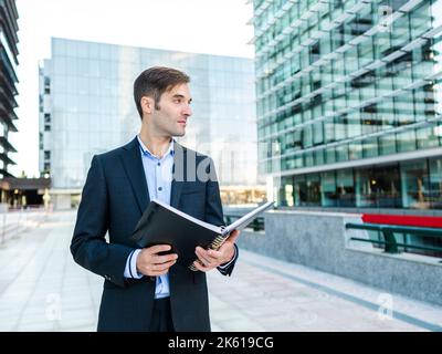 Male entrepreneur in elegant suit holding notepad and looking away dreamily on sunny street against contemporary building Stock Photo