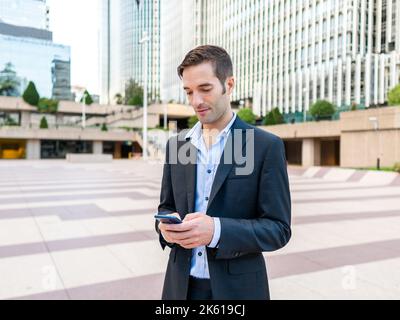 Young elegant man standing on pavement in modern city district and browsing mobile phone on sunny day Stock Photo
