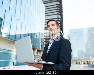 Low angle thoughtful entrepreneur in formal wear looking away while standing on city street and working on laptop Stock Photo