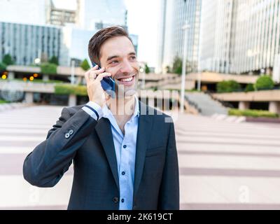 Young elegant man looking away while standing on pavement in modern city district and talking phone on sunny day Stock Photo