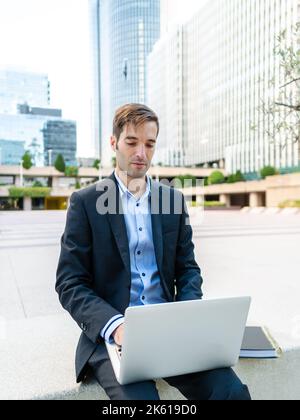 Concentrated guy in elegant suit sitting on bench in modern city and browsing computer while working on project in sunlight Stock Photo