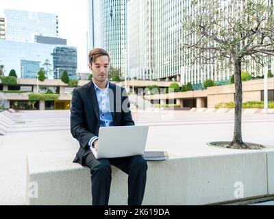 Concentrated guy in elegant suit sitting on bench in modern city and browsing computer while working on project in sunlight Stock Photo