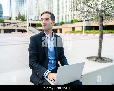 Concentrated guy in elegant suit sitting on bench in modern city with computer while working on project in sunlight Stock Photo