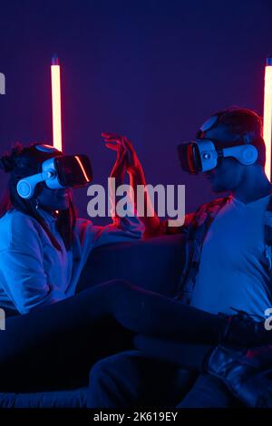 Delightful anonymous male and ethnic girlfriend in VR glasses sitting on sofa in darkness and connecting hands Stock Photo