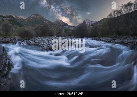 Fast stream of clean water flowing on rocks near mountain ridge covered with forest trees under starry sky at autumn night Stock Photo