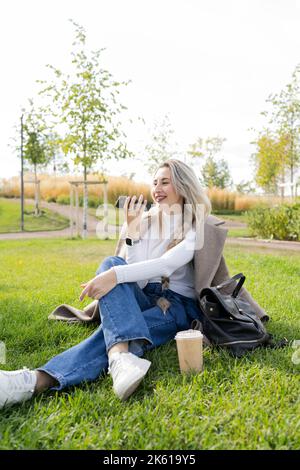 Cheerful female in stylish clothes smiling and recording audio message on cellphone while sitting on lawn near takeaway beverage and bag in park Stock Photo