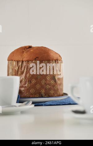 Uncut fresh baked artisan Christmas panettone cake on plate placed on blue cloth next to cups against white wall Stock Photo