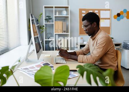 Side view portrait of young black man using smartphone at workplace in minimal office setting with plants Stock Photo