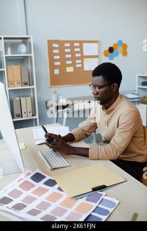 Vertical side view portrait of creative male designer using smartphone at workplace in office and working with color palettes for mobile app Stock Photo