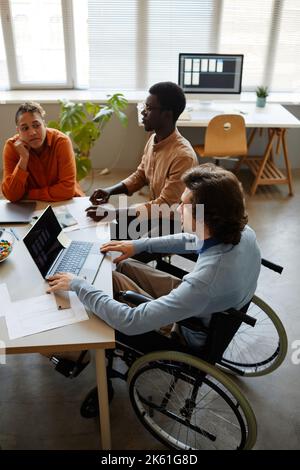 Vertical portrait of diverse business team discussing project at meeting table focus on young man using wheelchair in foreground Stock Photo