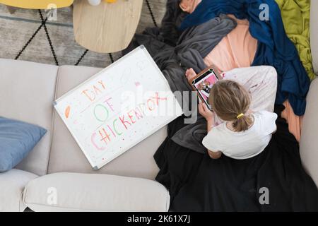 Top view of Little school girl with chickenpox playing creative games on tablet and resting on sofa at home, antiseptic cream applied to face and body Stock Photo