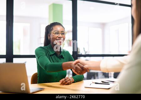 Happy female coworkers shaking hands in the office, african-american businesswoman smiling. Teamwork, partnerships and hiring concept Stock Photo