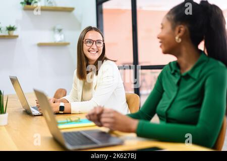 Two diverse female employees in smart casual wear working with laptops together in the contemporary meeting room, sharing plans and ideas with each other in friendly working atmosphere Stock Photo