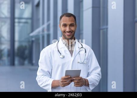 Portrait of happy and smiling hispanic doctor, man smiling and looking at camera, doctor using tablet computer for online consultation, outside of modern clinic. Stock Photo