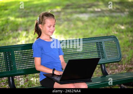 Schoolgirl studying on laptop computer resting on park bench outdoors on summer day. Remote education during quarantine concept Stock Photo