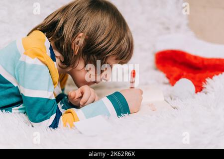 Smiling Kid boy lying on couch and writing the letter to dear Santa at home in Christmas holidays. Child wish list. Dreams of a Christmas gifts. Merry Stock Photo