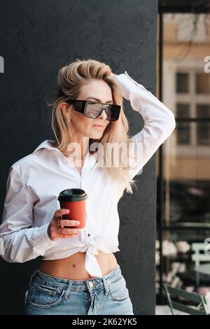 Elegant Blonde Woman Holding Cup of Coffee in Hand and Posing near the Wall on Terrace of Cafe, Confident and Fashionable Female Model Wearing Stock Photo