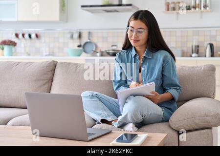 Teenager asian woman watching webinar on laptop making notes Stock Photo