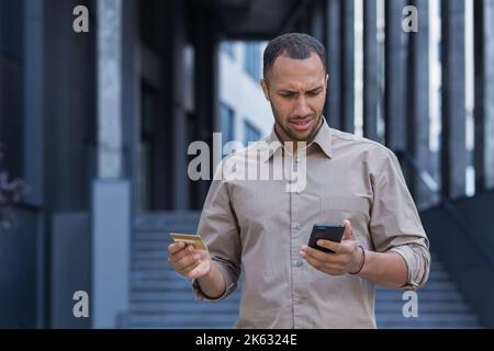 Frustrated and upset african american man outside office building trying to make a purchase in an online store, man using smartphone and bank credit card, to transfer money and buy online. Stock Photo