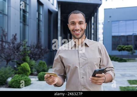 Portrait of happy hispanic man outside office building looking at camera and smiling, businessman holding phone and bank credit card for online shopping. Stock Photo