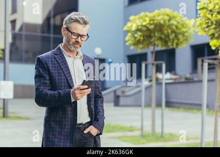 Serious and focused mature businessman using smartphone outside office building, gray haired man in glasses and business suit reading online message, senior boss walking in business city. Stock Photo
