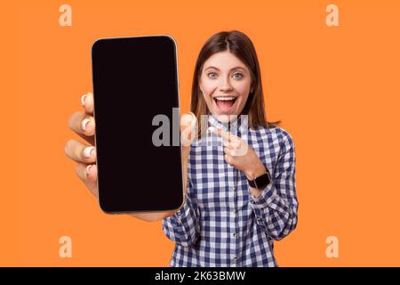 Portrait of delighted amazed young woman wearing checkered shirt pointing at smart phone with empty display, copy space for advertisement. Indoor studio shot isolated on orange background. Stock Photo