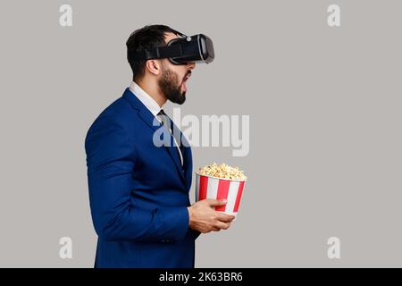 Side view of excited amazed bearded businessman in VR headset watching movie with popcorn, keeps mouth open, wearing official style suit. Indoor studio shot isolated on gray background. Stock Photo