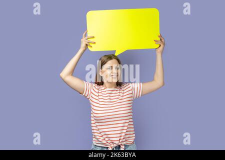 Portrait of positive smiling satisfied blond woman wearing striped T-shirt holding yellow bubble for text, looking at camera with toothy smile. Indoor studio shot isolated on purple background. Stock Photo