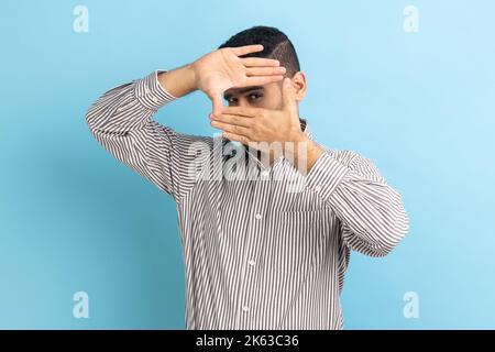 Creative businessman looking through hand frame gesture, cropping image, zooming and focusing on distance, viewing target, wearing striped shirt. Indoor studio shot isolated on blue background. Stock Photo
