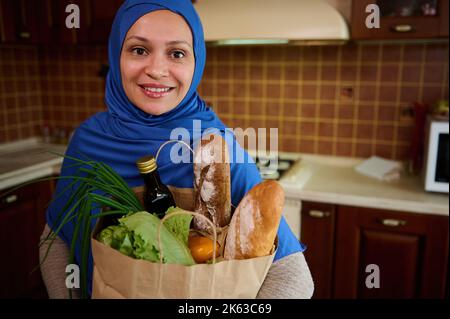 Young Arab Muslim woman with head covered in blue hijab, unpacking shopping bag with organic food on the kitchen at home Stock Photo