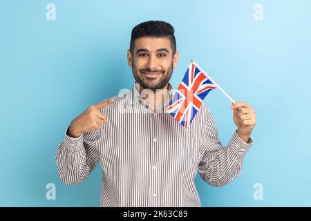 Portrait of satisfied businessman with beard standing pointing at British flag, looking at camera, patriot, wearing striped shirt. Indoor studio shot isolated on blue background. Stock Photo