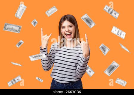 Portrait of excited woman wearing striped shirt standing with rock sign and shouting for joy, amazed of money rain falling from up. Indoor studio shot isolated on orange background. Stock Photo