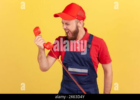 Portrait of angry aggressive worker wearing blue overalls screaming to client on handset, does not accept order, incompetent service personnel. Indoor studio shot isolated on yellow background. Stock Photo