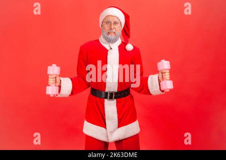 Portrait of strong elderly man with gray beard wearing santa claus costume standing with dumbbells in hands, trying to lift up heavy sport equipment. Indoor studio shot isolated on red background. Stock Photo